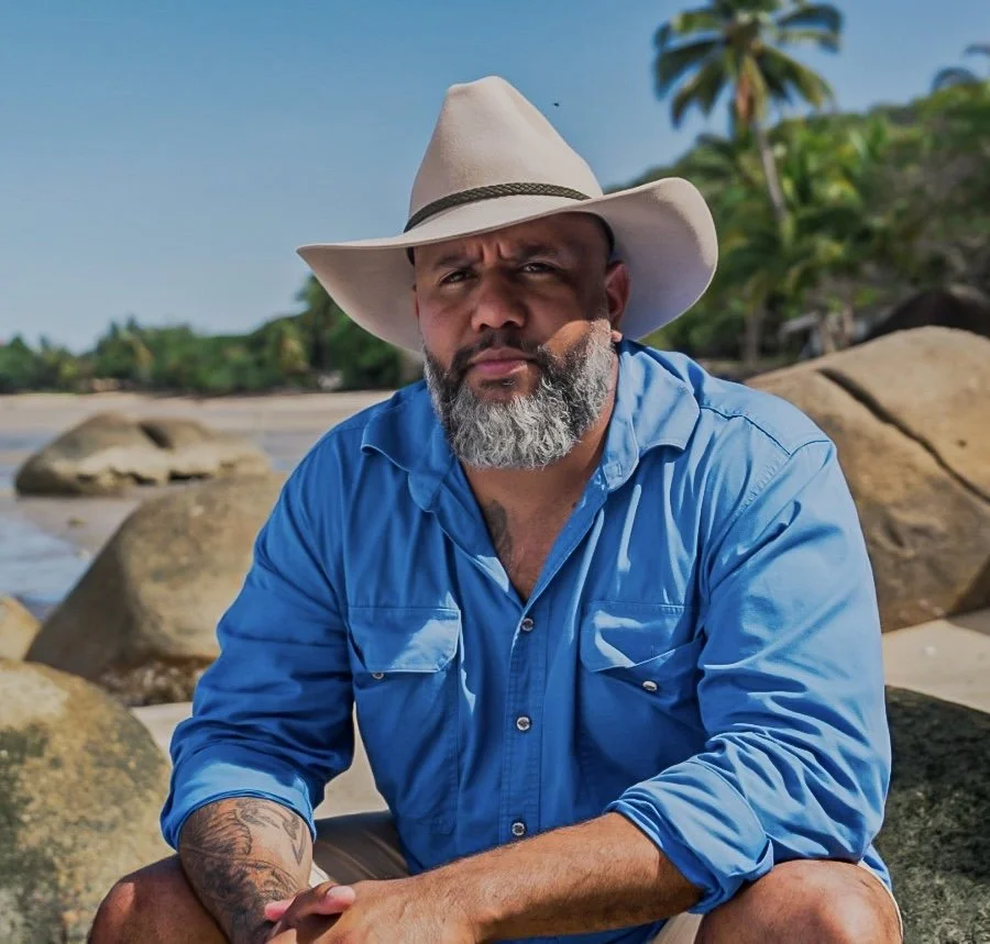 A man with a gray beard wearing a wide-brimmed hat and a blue shirt, sitting on rocks at a tropical beach with palm trees in the background.