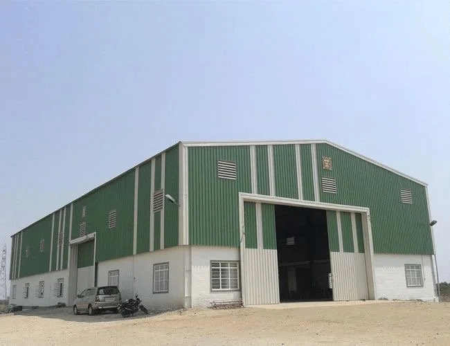 Large industrial warehouse with green metal upper walls and white lower walls, multiple ventilation openings, and an open garage door, with vehicles parked nearby
