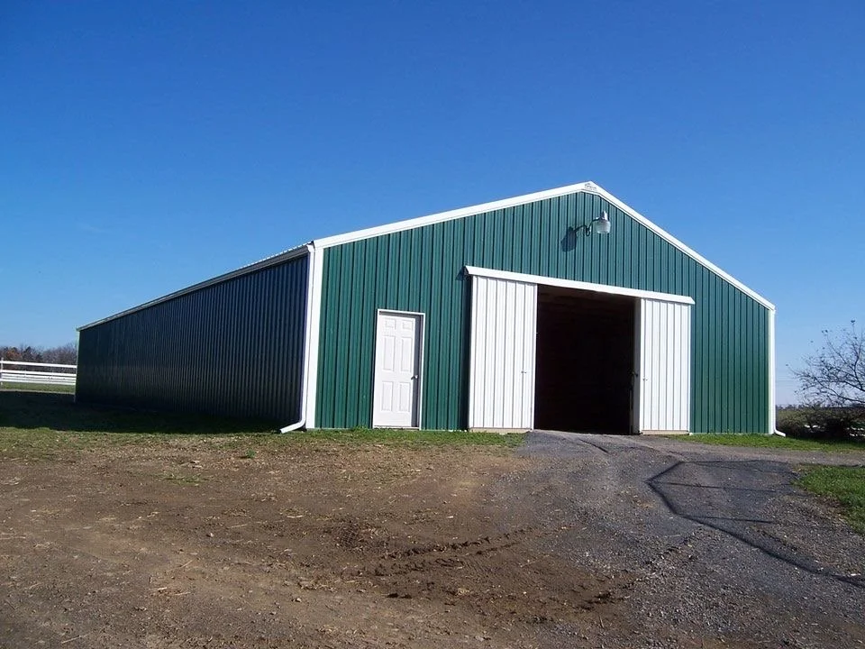 Green metal barn with white trim, white door, and open garage door on a clear day with blue sky.
