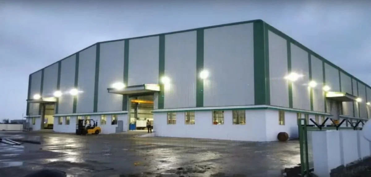 Large commercial warehouse building with green and white exterior, side lighting, and a loading dock area in a wet, overcast environment.