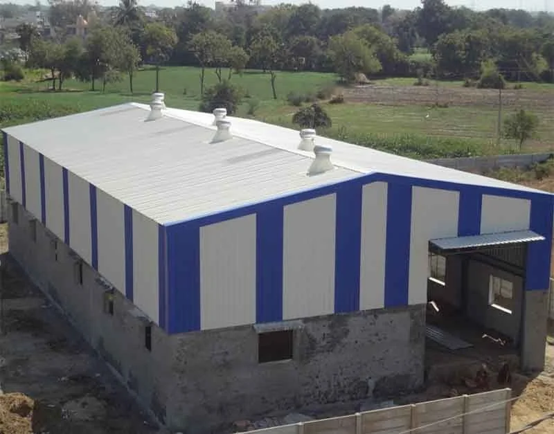 A large industrial warehouse with blue and white vertical striped walls and a metal roof, situated in a rural area with green fields and trees in the background.