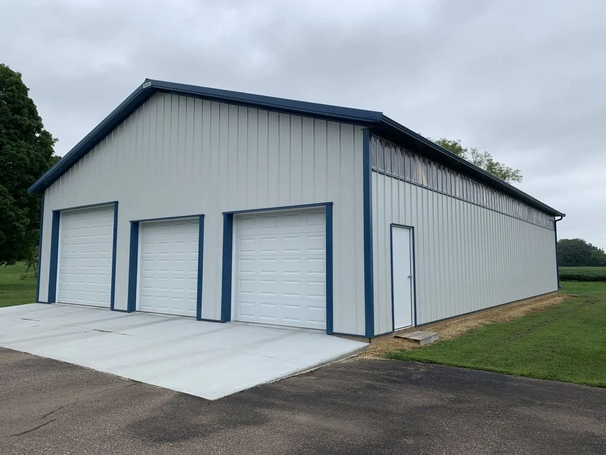 Large metal storage building with three garage doors and a side access door, situated on a concrete and asphalt surface with green grass and trees nearby.