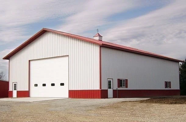 Large white barn with red trim and roof, featuring a big garage door, small windows, and a gravel ground, under a partly cloudy sky.