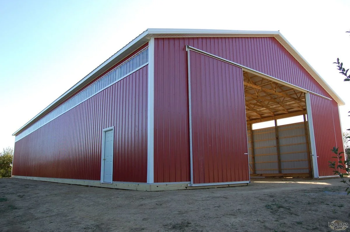 A red metal building with an open section showing the wooden frame inside, situated on dirt ground with a small door on the side.