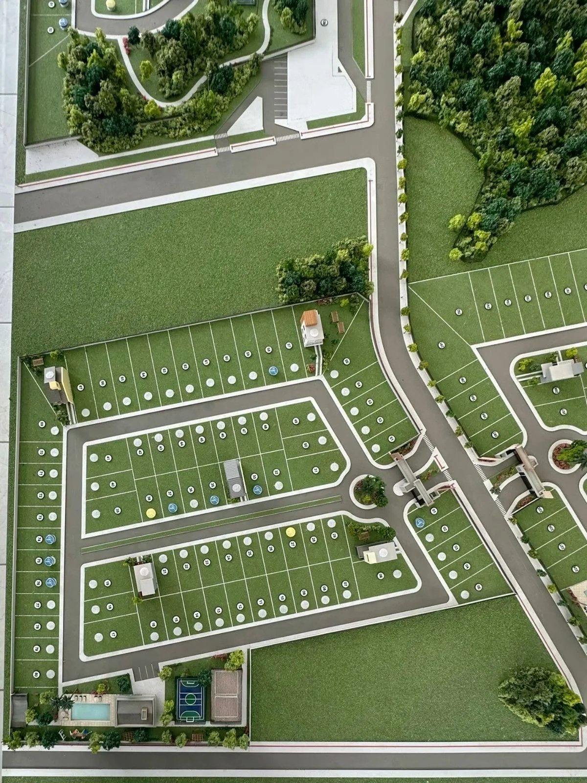 Aerial view of an apartment complex with parking lots, green lawns, trees, and wooded areas, including a swimming pool and sports court.