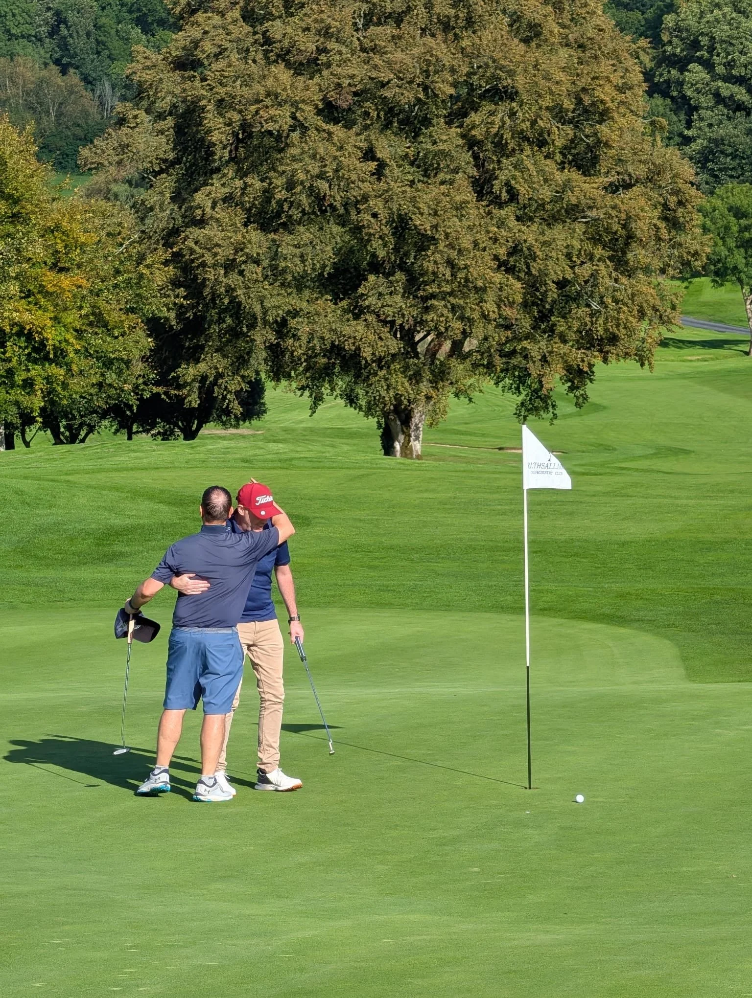 Two men embrace on a golf course near a putting green with a golf flag and a golf ball, large trees in the background, and bright sunny weather.