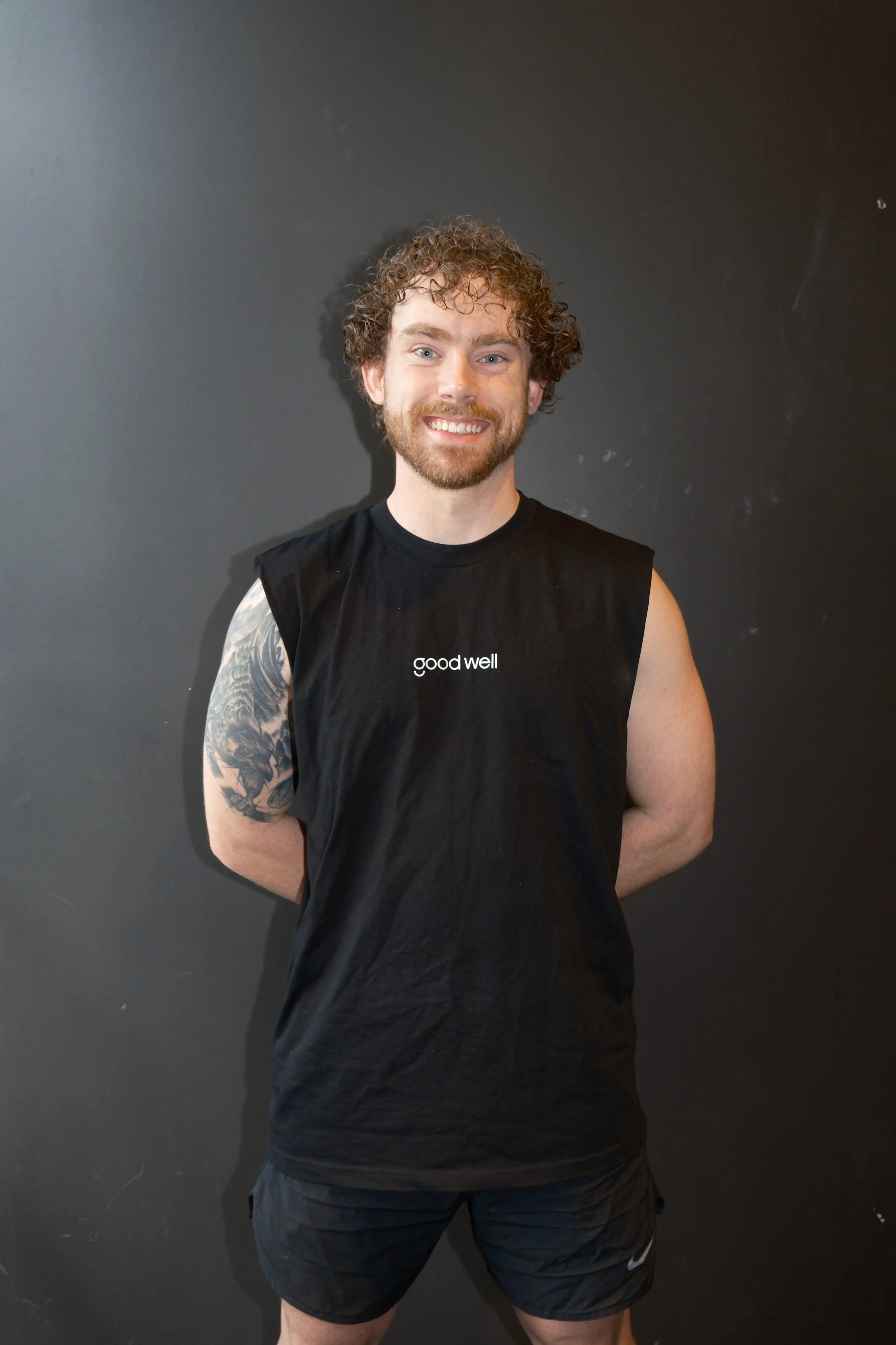 Young man with curly hair and beard smiling, wearing a black t-shirt with a small Good Well logo, sitting outdoors in front of a the gym.