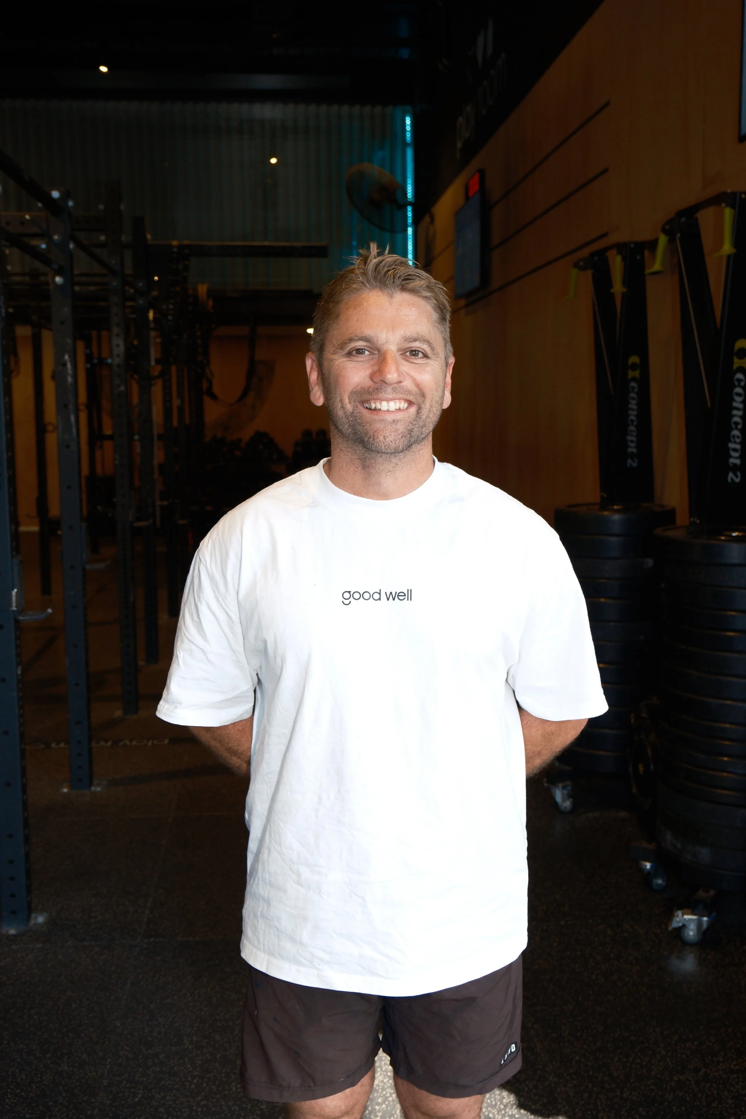 A smiling man in a sleeveless black shirt with 'good well' written on it, standing in front of a white wall with a backlit sign.