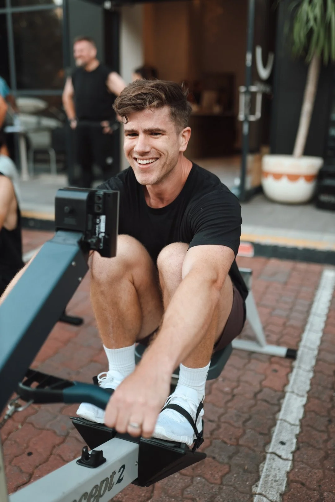 A smiling man with brown hair and a beard sitting on a couch in a white t-shirt with a good well shirt