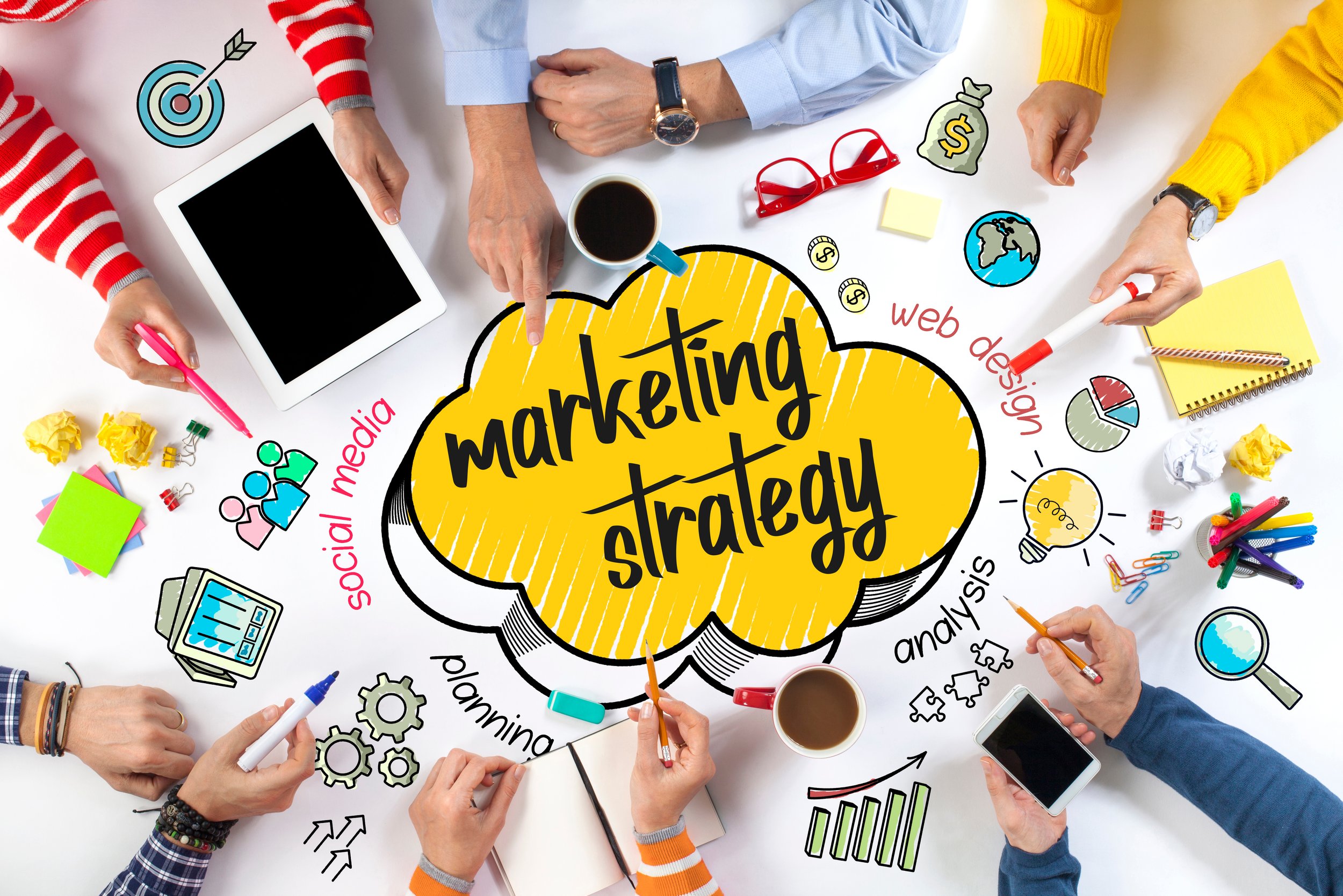 A group of people working together on a marketing strategy, surrounded by electronic devices, coffee cups, and colorful stationery. The central focus is a yellow cloud-shaped graphic with the words "marketing strategy" and related terms like social media, analysis, and web design written around it.
