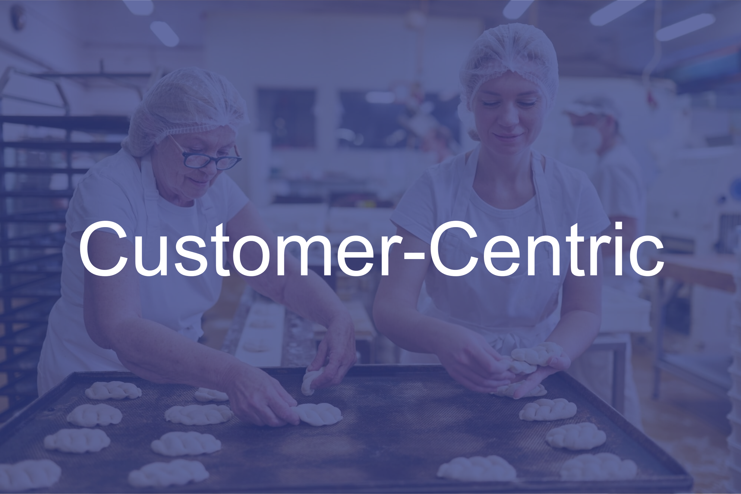 Two women baking bread dough in a commercial kitchen, dressed in white uniforms and hairnets, with the word "Customer-Centric" displayed in the foreground.