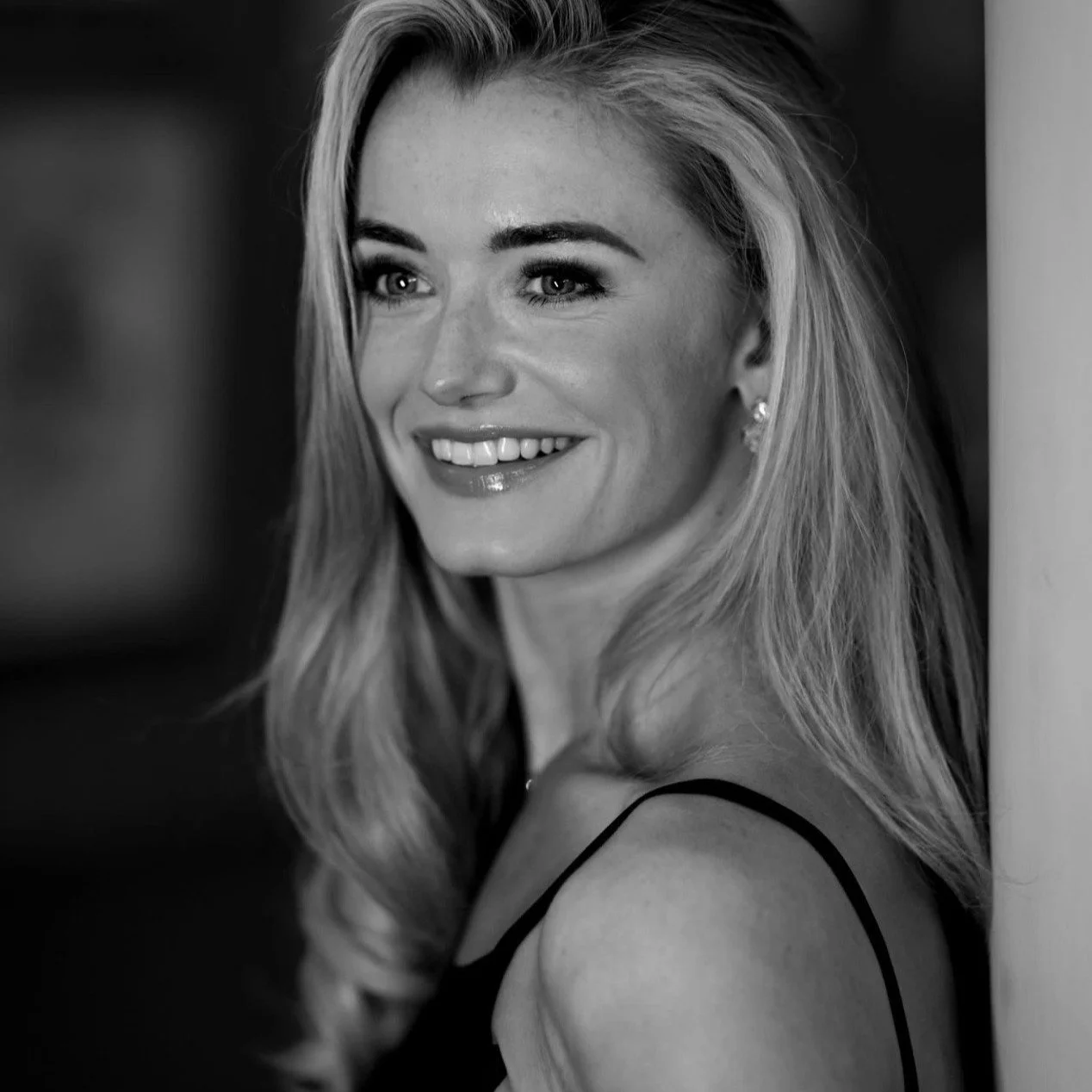 Anna Rose O'Sullivan, smiling with wavy blonde hair, wearing a sleeveless top, standing near a pillar.