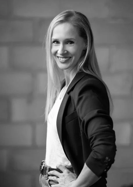Black and white photo of a smiling young woman with long straight hair, wearing a blazer over a white shirt, standing with her hand on her hip in front of a brick wall.