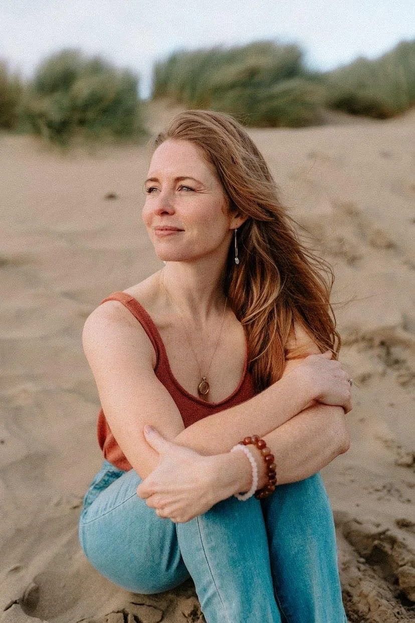 Rachael Hall is sitting on sandy ground at the beach, arms crossed over her knees, gazing into the distance, wearing a rust-colored tank top, light blue jeans, and jewellery, including necklaces, earrings, and bracelets.