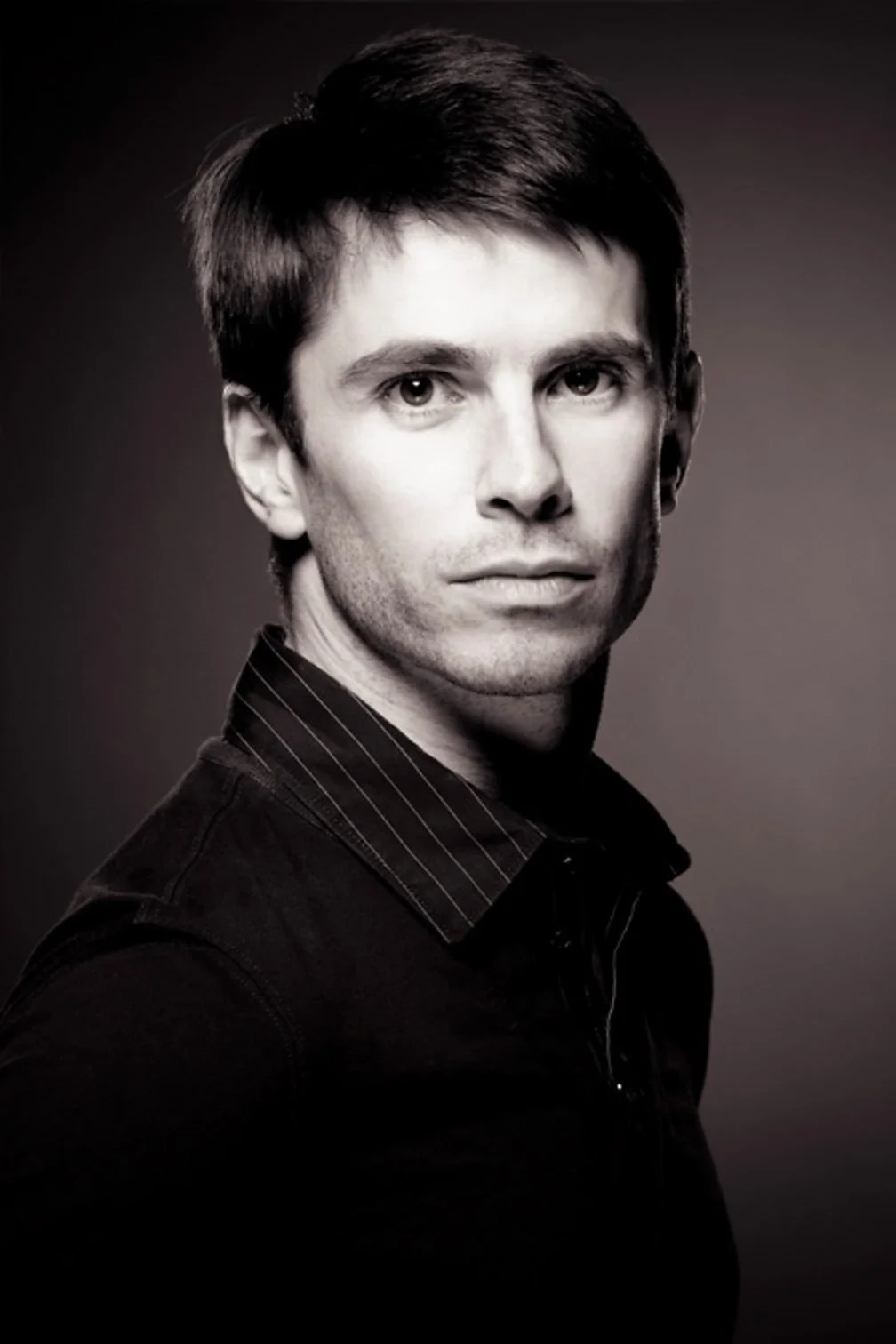 Black and white portrait of Ricardo Cervera wearing a dark shirt with thin vertical stripes, looking directly at the camera with a serious expression.