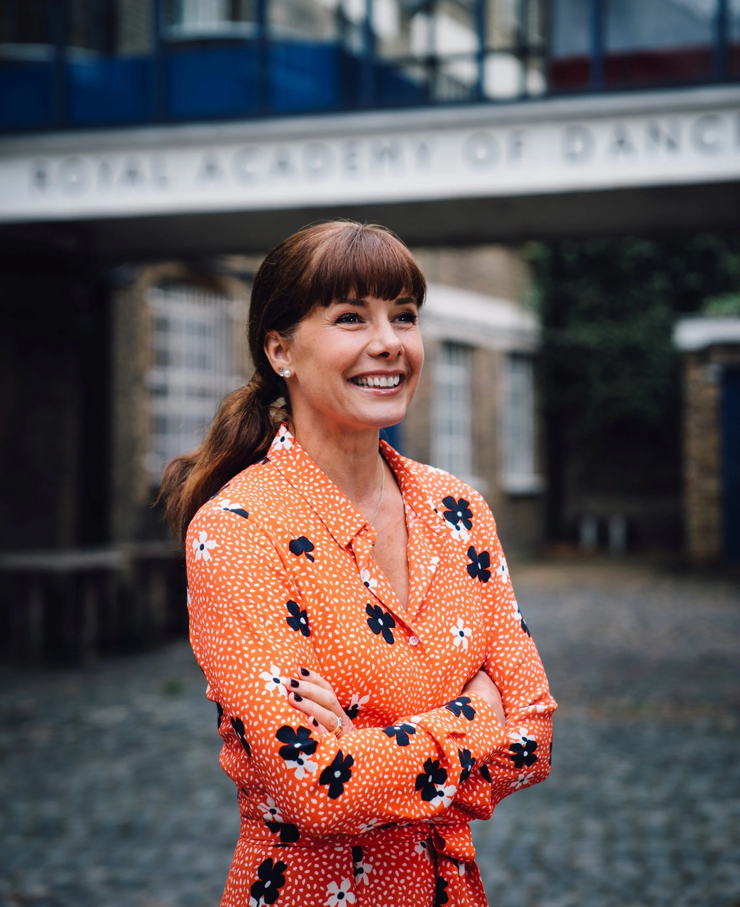Dame Darcey Bussell, wearing a bright orange shirt with black, white, and red floral patterns, smiles outdoors in front of a building with a sign that reads "Royal Academy of Dance."