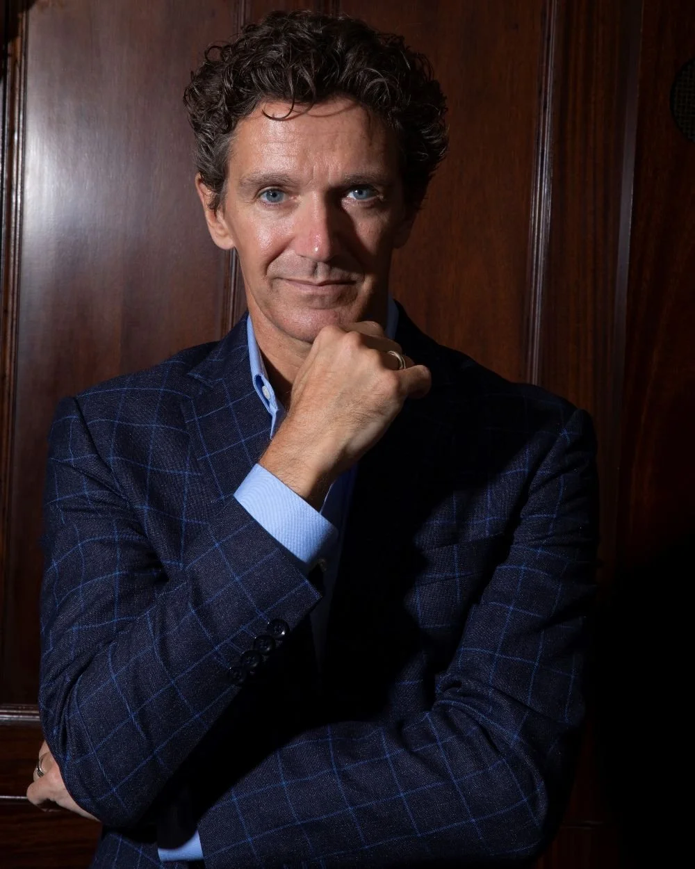 A picture of Gary Avis MBE, wearing a dark plaid blazer and a light blue shirt, standing against a wooden wall. He is posing with his right hand under his chin and smiling lightly.