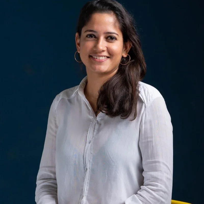 A woman with long dark hair, smiling, wearing a white shirt and hoop earrings, standing against a dark background.