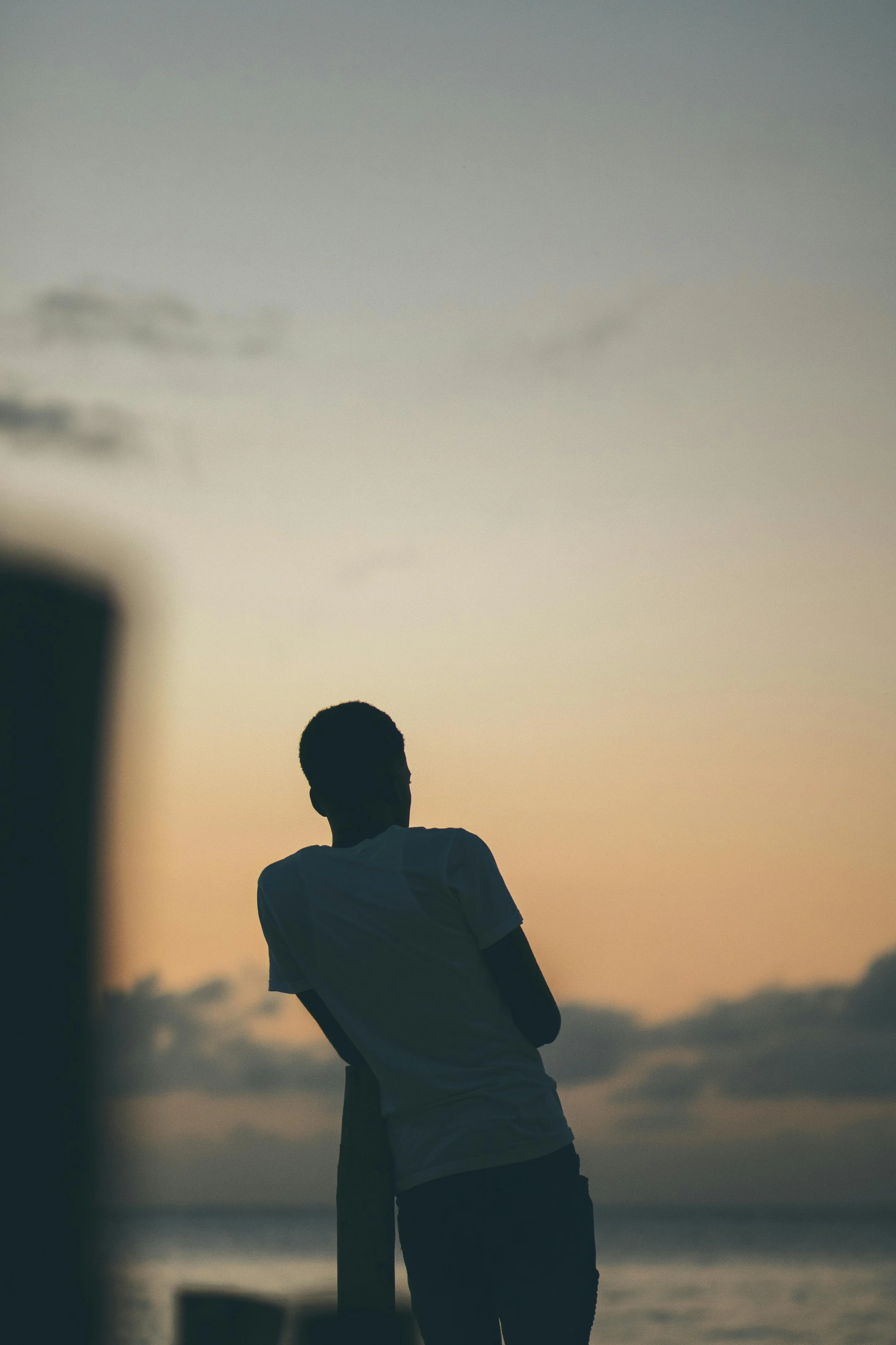 Silhouette of a young man leaning against a post and looking at the sunset or sunrise sky with clouds.