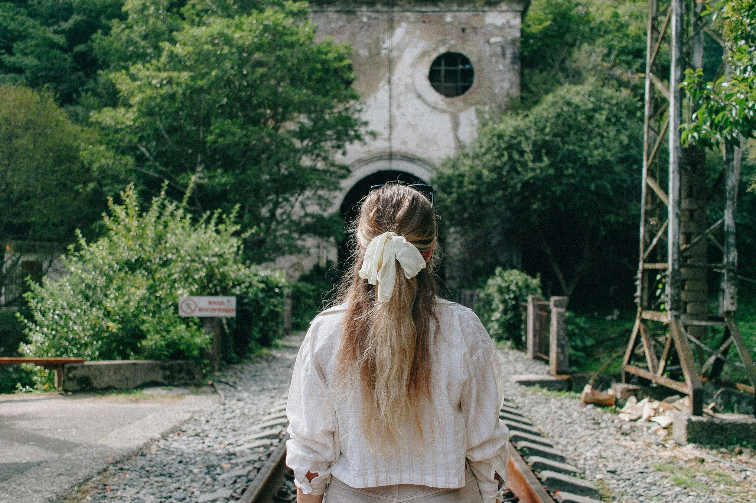 A woman with long, blonde hair tied with a cream-colored bow, standing on a train track facing away, surrounded by green trees and an old building in the background.