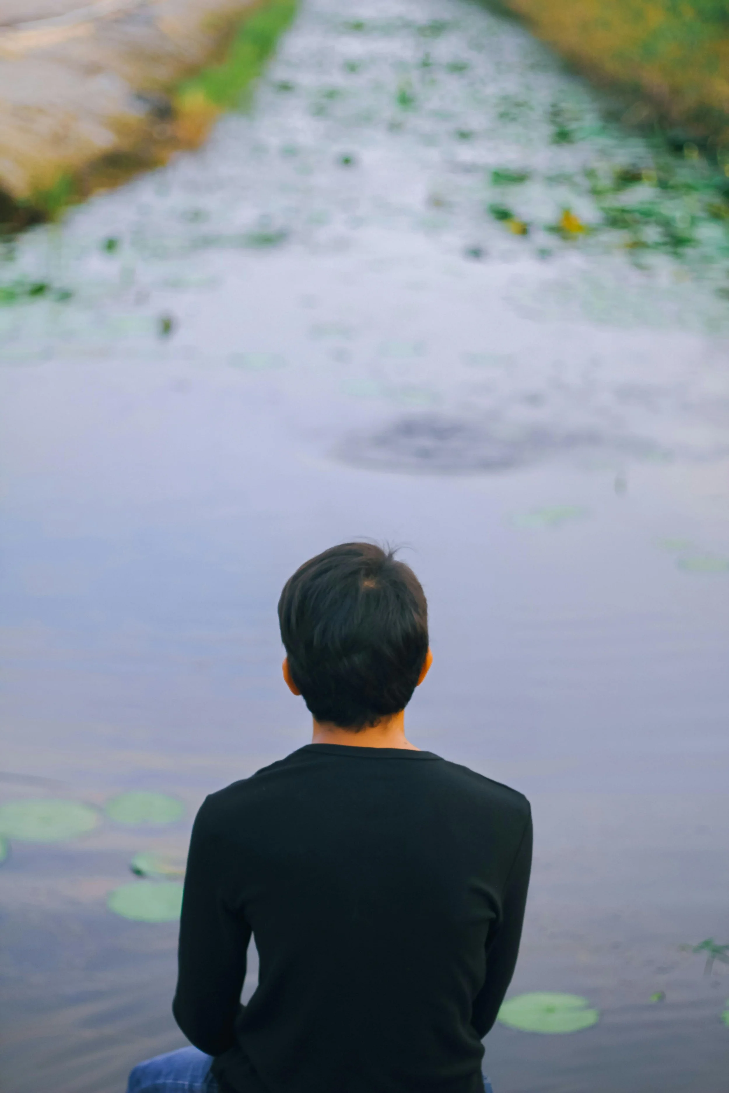 A person with dark hair wearing a black shirt sitting by a pond with floating lily pads, looking away from the camera.