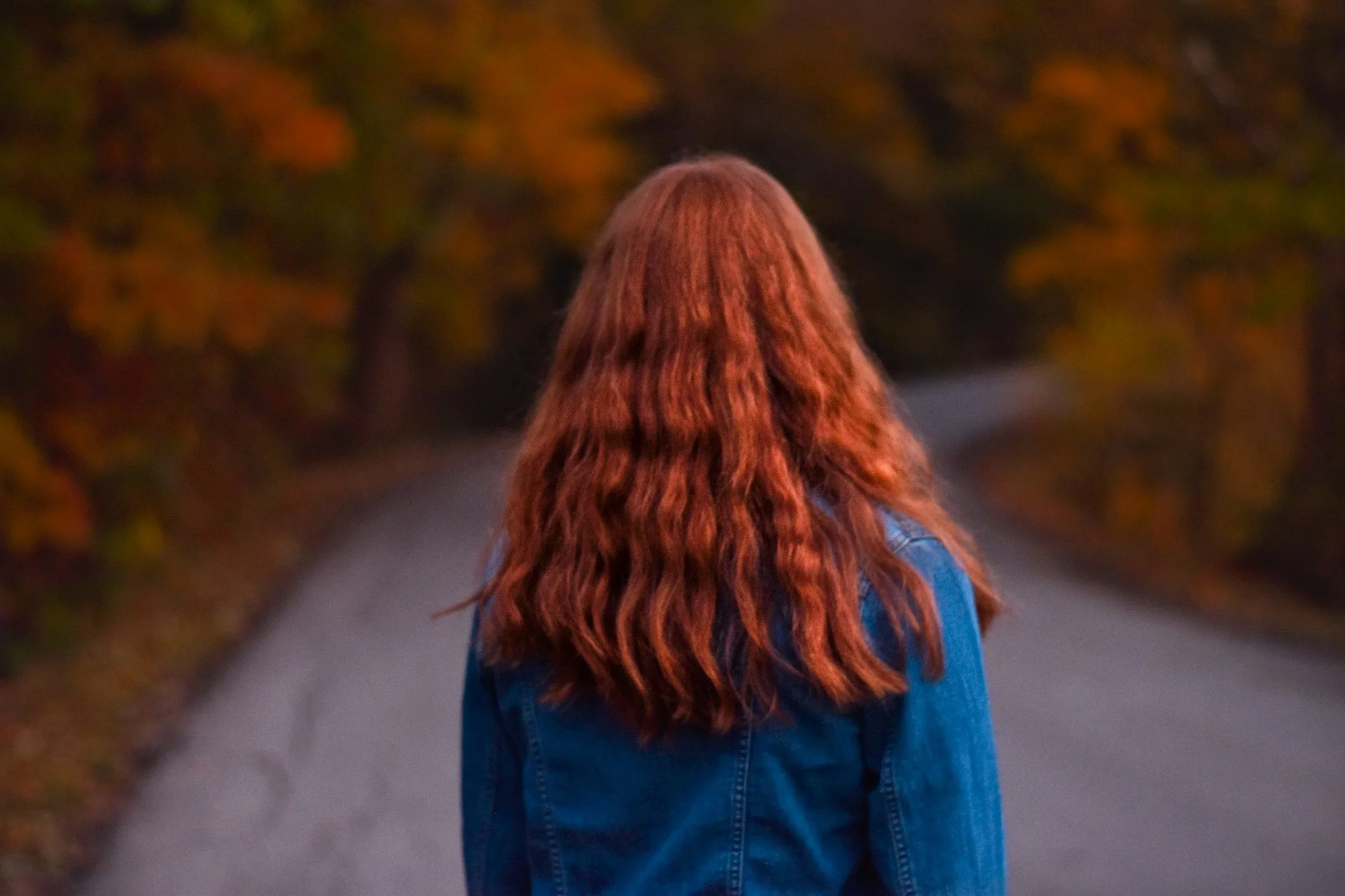 A person with long wavy red hair wearing a denim jacket, facing away on a winding road surrounded by autumn trees.
