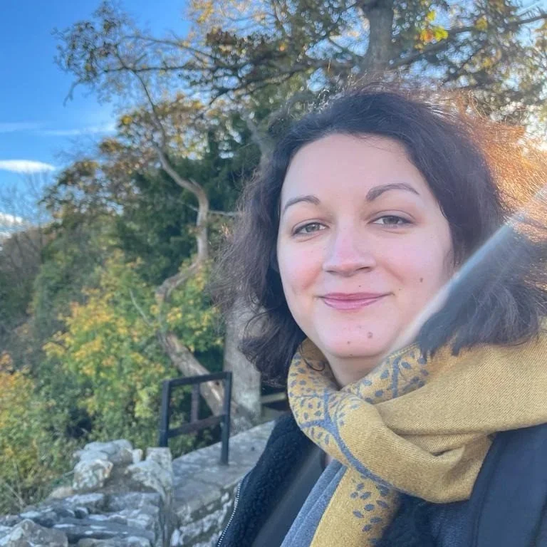 A woman with dark curly hair smiling outdoors, wearing a yellow scarf with a floral pattern and a dark jacket, with trees and a rocky area in the background.