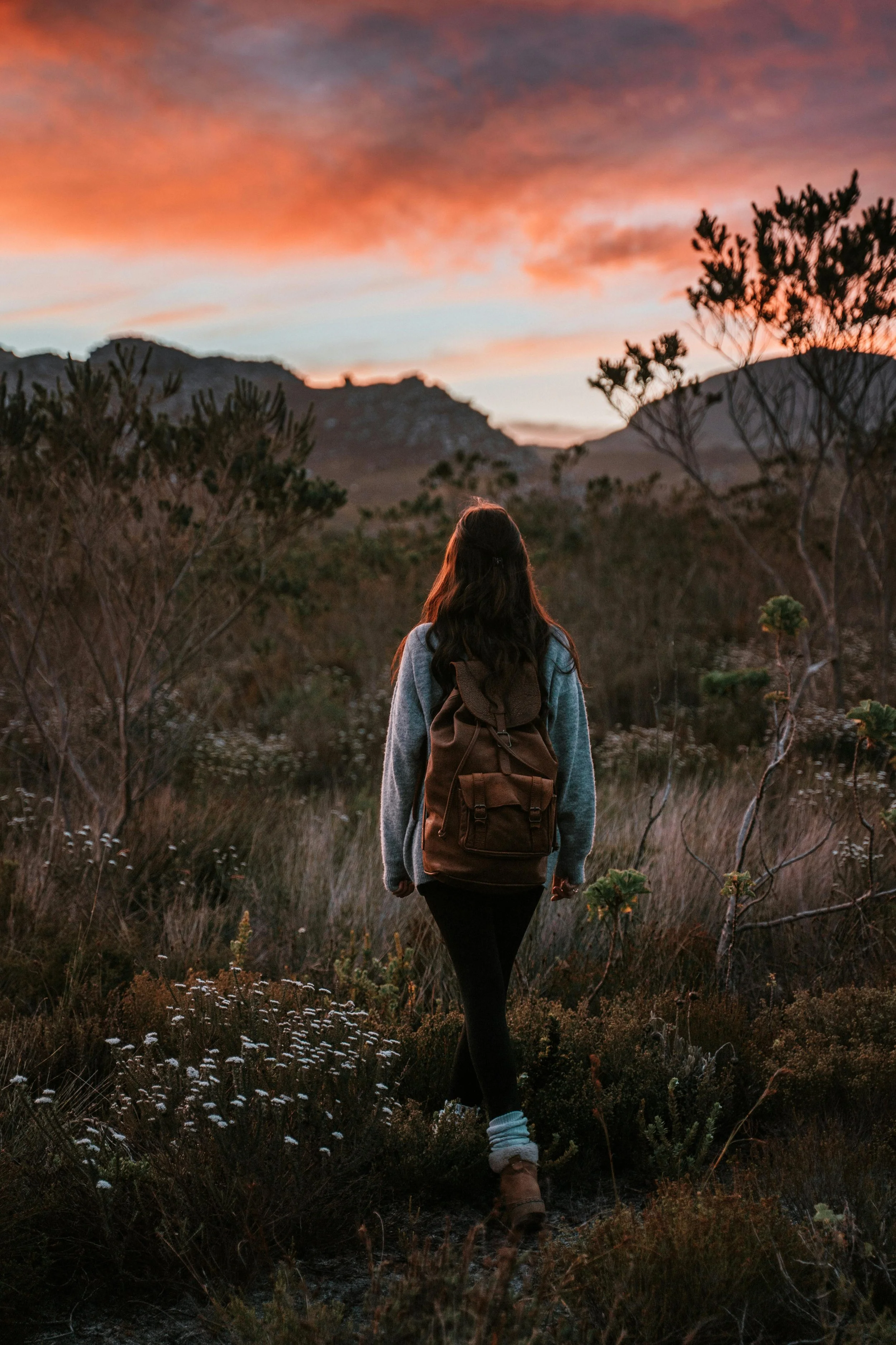 A woman with long hair, wearing a backpack and boots, walking through a dry, grassy landscape with bushes at sunset, with colorful clouds and mountain silhouettes in the background.