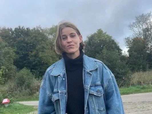 A young woman standing outdoors with trees in the background, wearing a black turtleneck and a blue denim jacket.