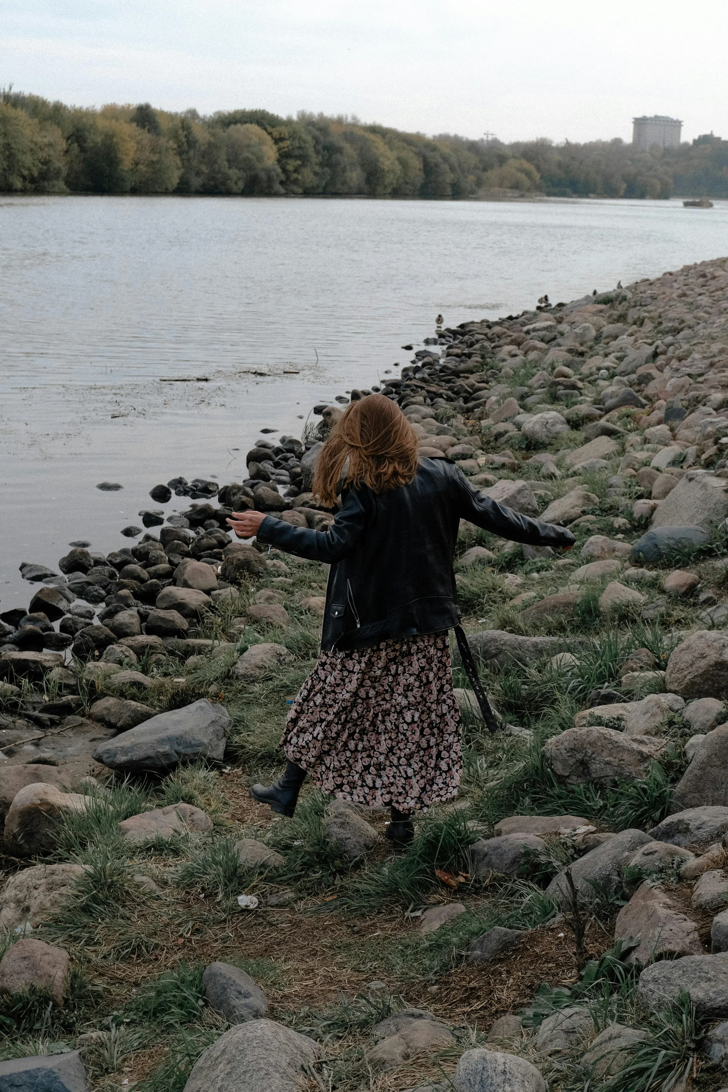 A woman with red hair, wearing a black leather jacket, floral skirt, and black boots, walking along a rocky riverside with trees and a large building in the background.