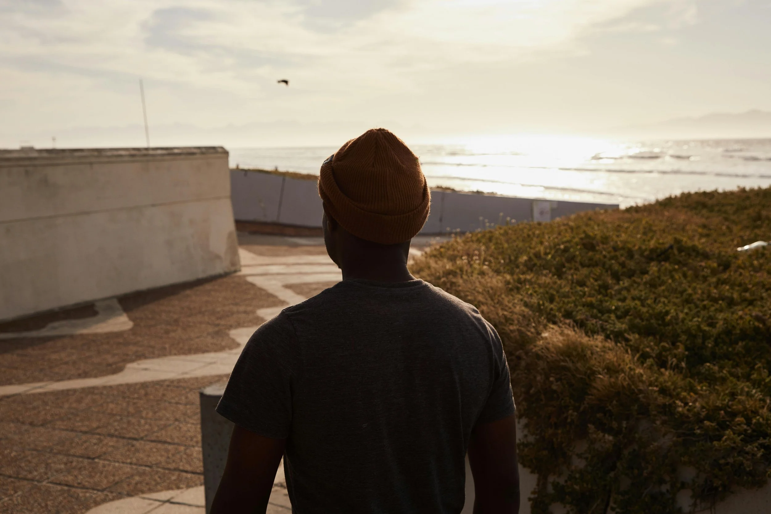 Person with a brown beanie and gray t-shirt standing outside near the ocean, facing the sunset over the water.