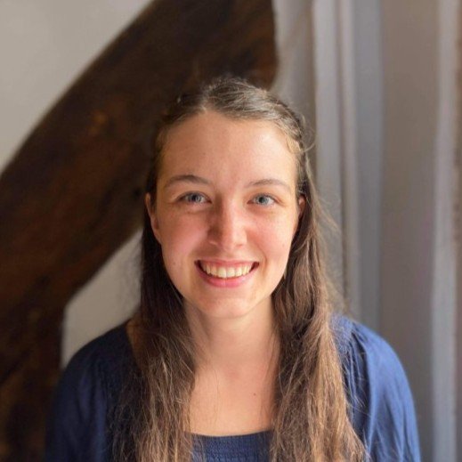 A young woman with long brown hair smiling at the camera, wearing a blue top, standing indoors near a window.