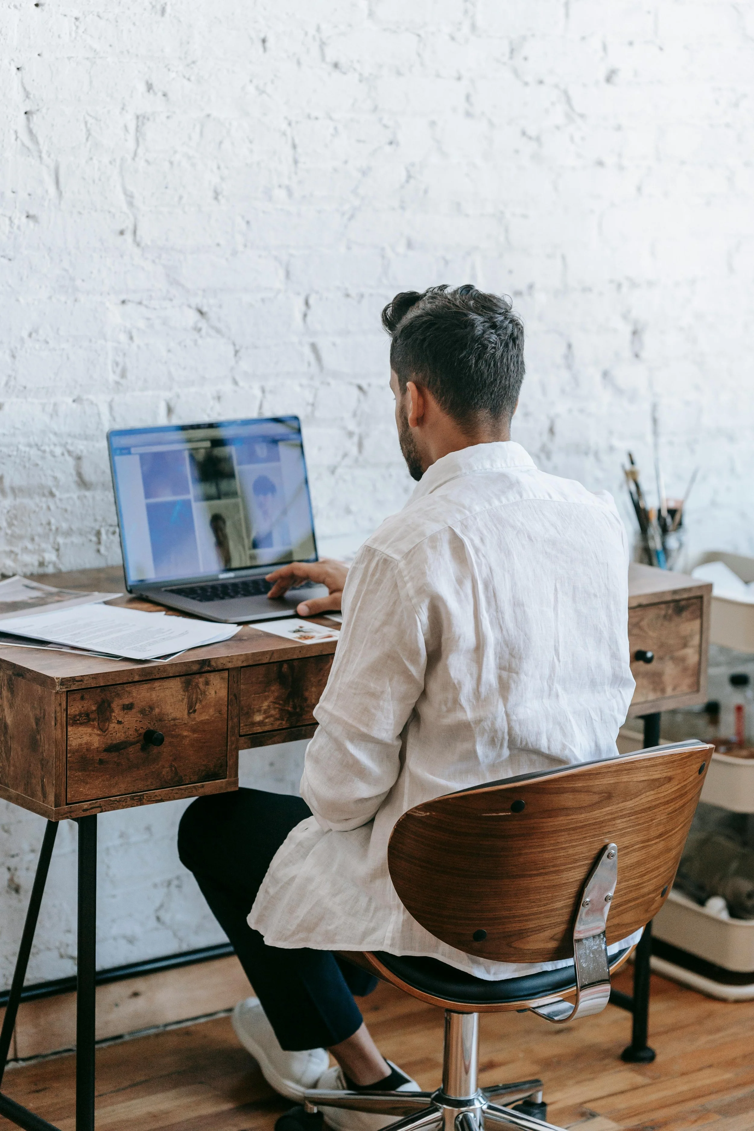 Man working on a laptop at a wooden desk in a room with a white brick wall.