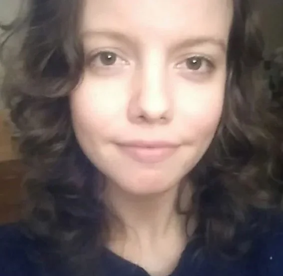Close-up of a woman with fair skin and curly dark hair, smiling slightly at the camera.
