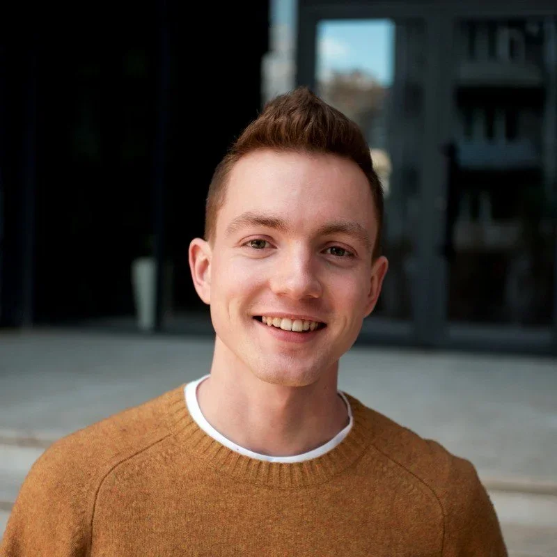 A young man with short, reddish-brown hair smiling outdoors in front of a modern building with glass windows.