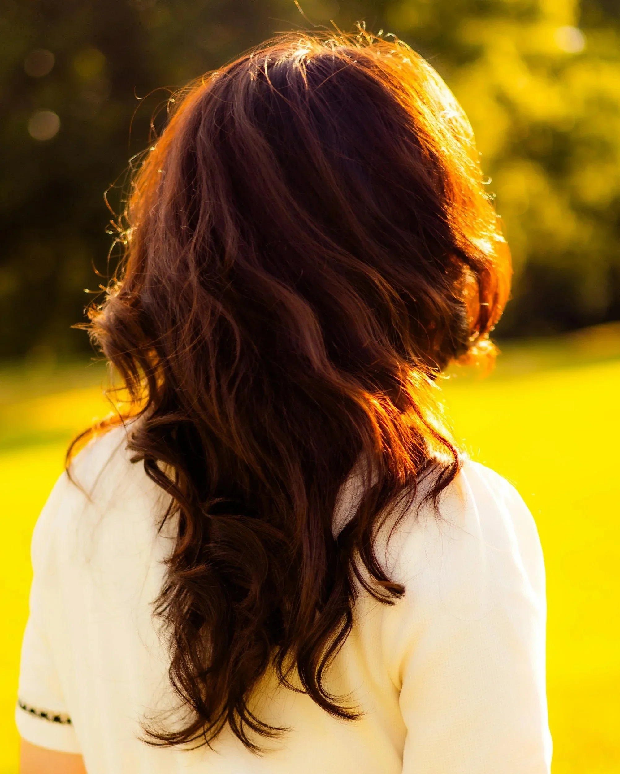 A woman with long, wavy brown hair standing outdoors in the sunlight, facing away from the camera, with a blurred green background.
