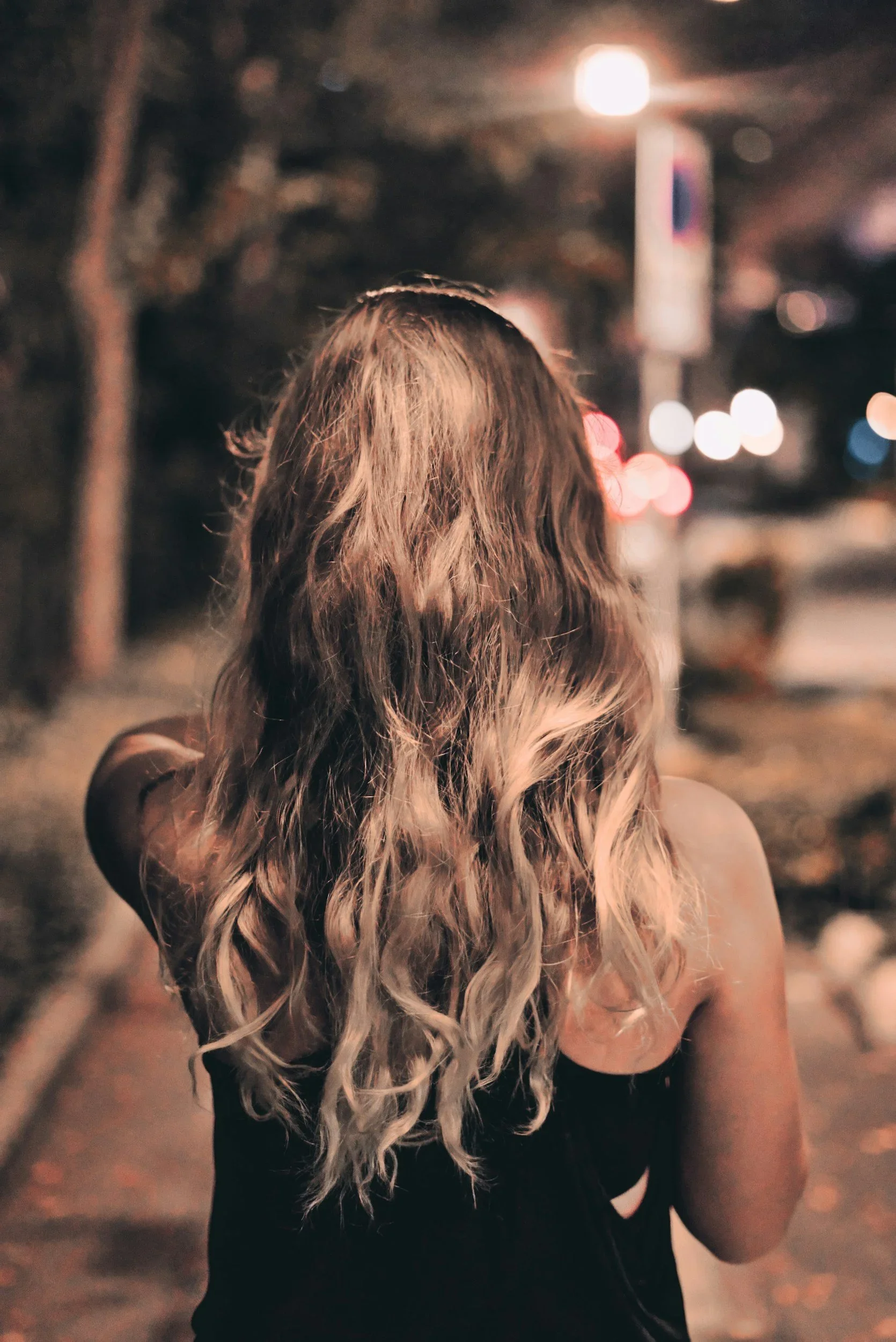 A woman with long, wavy blonde hair walking on a city sidewalk at night, seen from behind. Streetlights and blurred city lights are visible in the background.