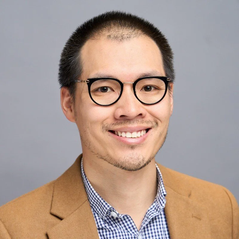 A man in his late 30s with glasses and a light brown blazer smiling at the camera.
