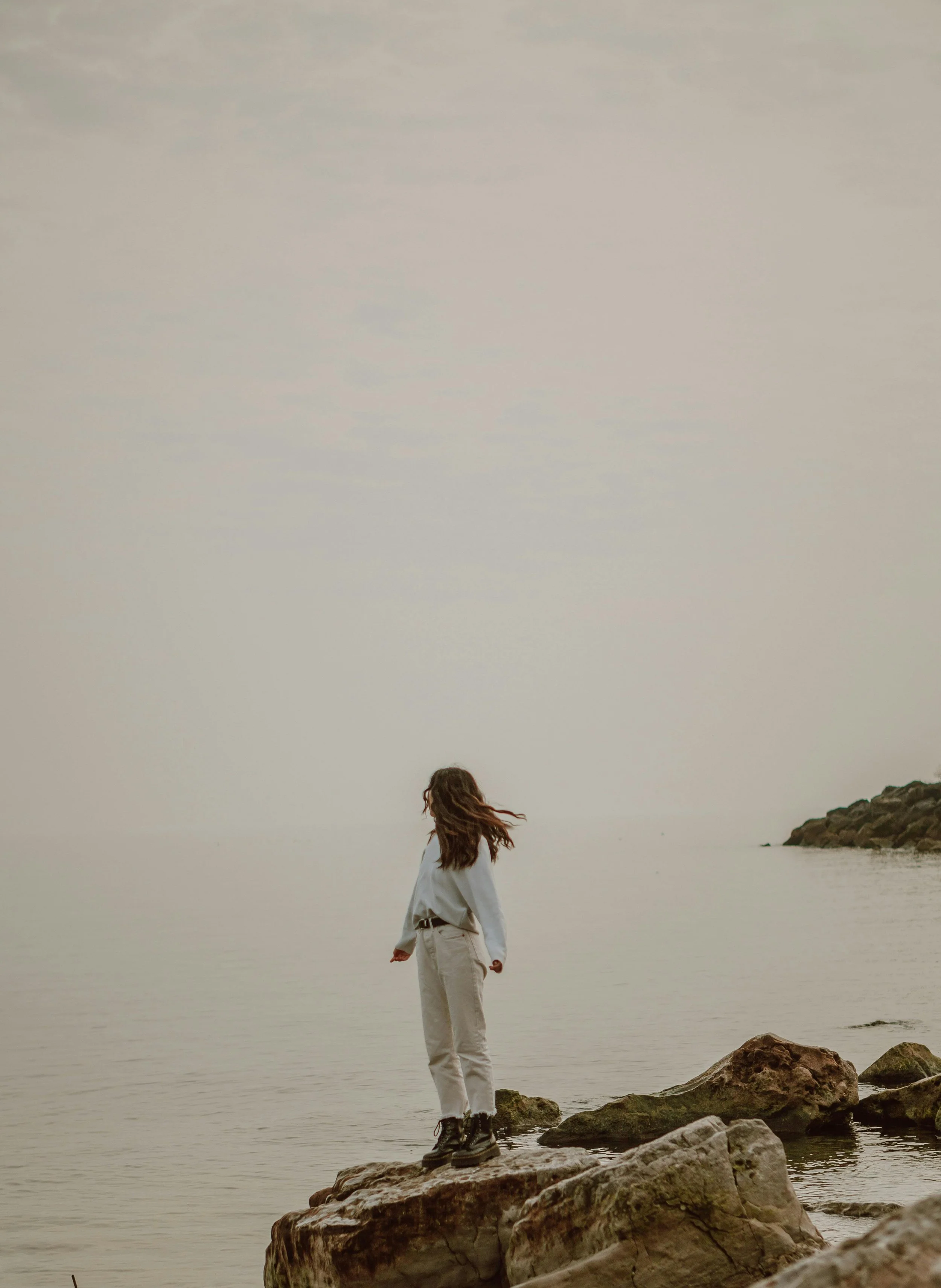 A person with long hair standing on rocks by the water's edge, facing the calm sea or lake with a cloudy sky above.