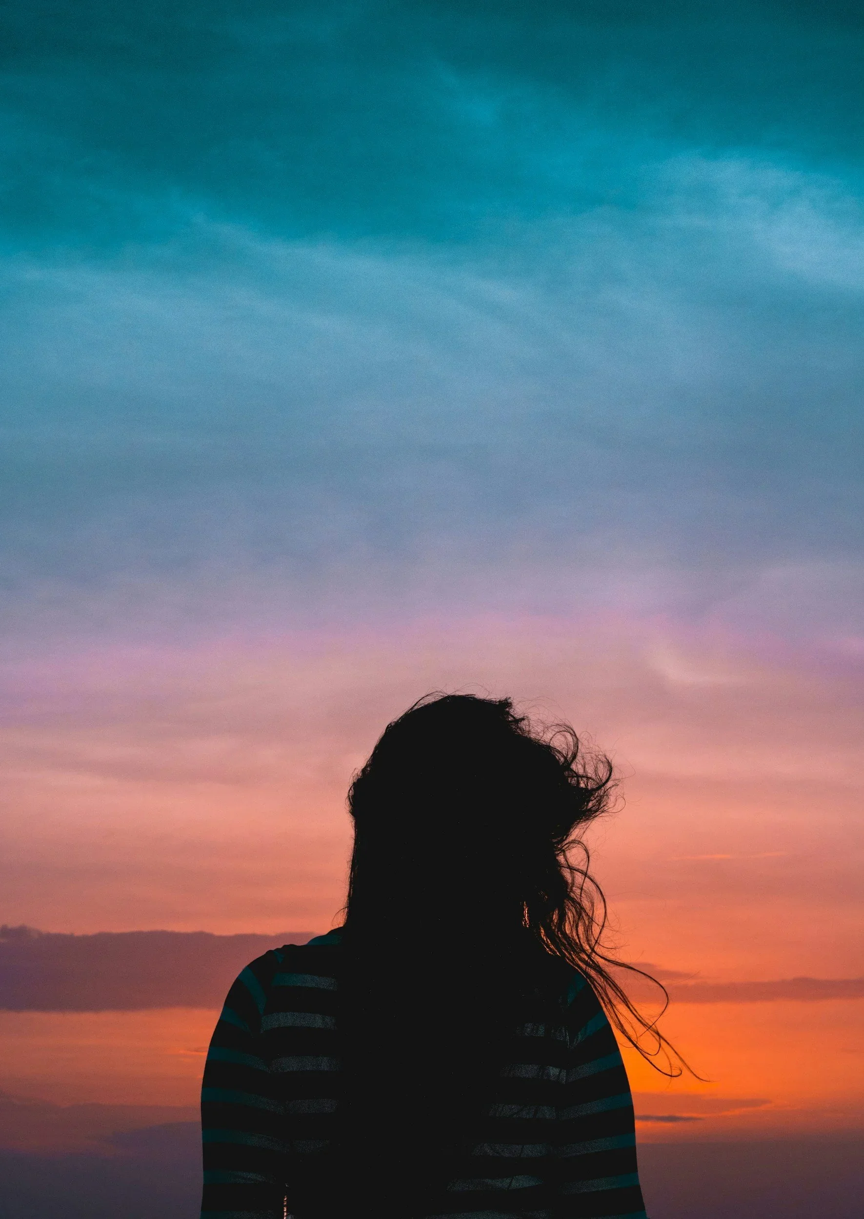 Silhouette of a person with long hair against a colorful sunset sky with blue, pink, purple, and orange hues.