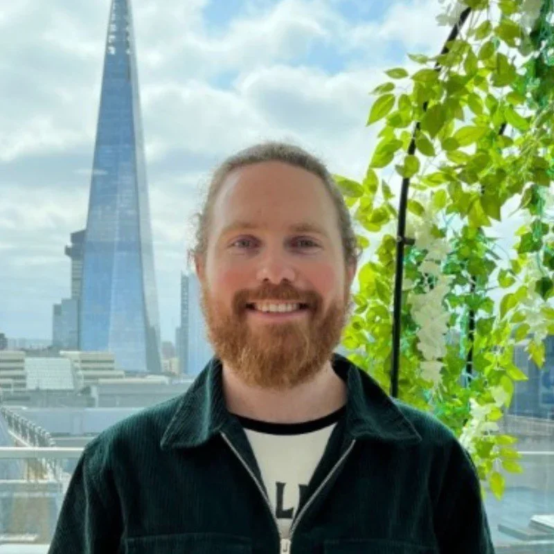 A smiling man with a beard and long hair standing on a rooftop with city buildings, including the Shard, in the background, and green foliage beside him.