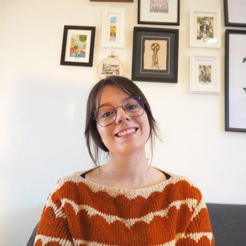 A young woman with glasses smiling, sitting indoors with a wall decorated with various framed artwork in the background.
