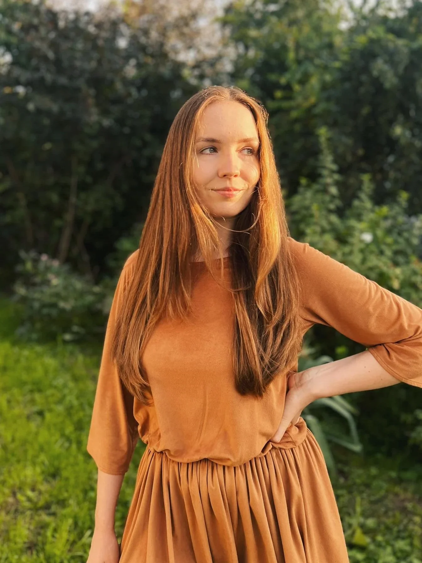 A young woman with long red hair standing outdoors with trees and greenery in the background. She is wearing a brown dress and has a slight smile on her face.