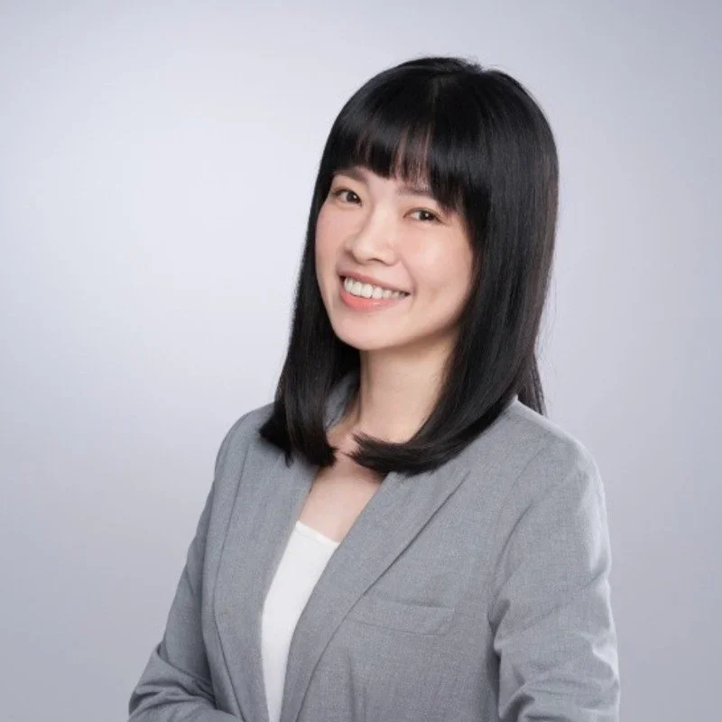 A woman with straight black hair and bangs, wearing a gray blazer and white shirt, smiling against a light gray background.