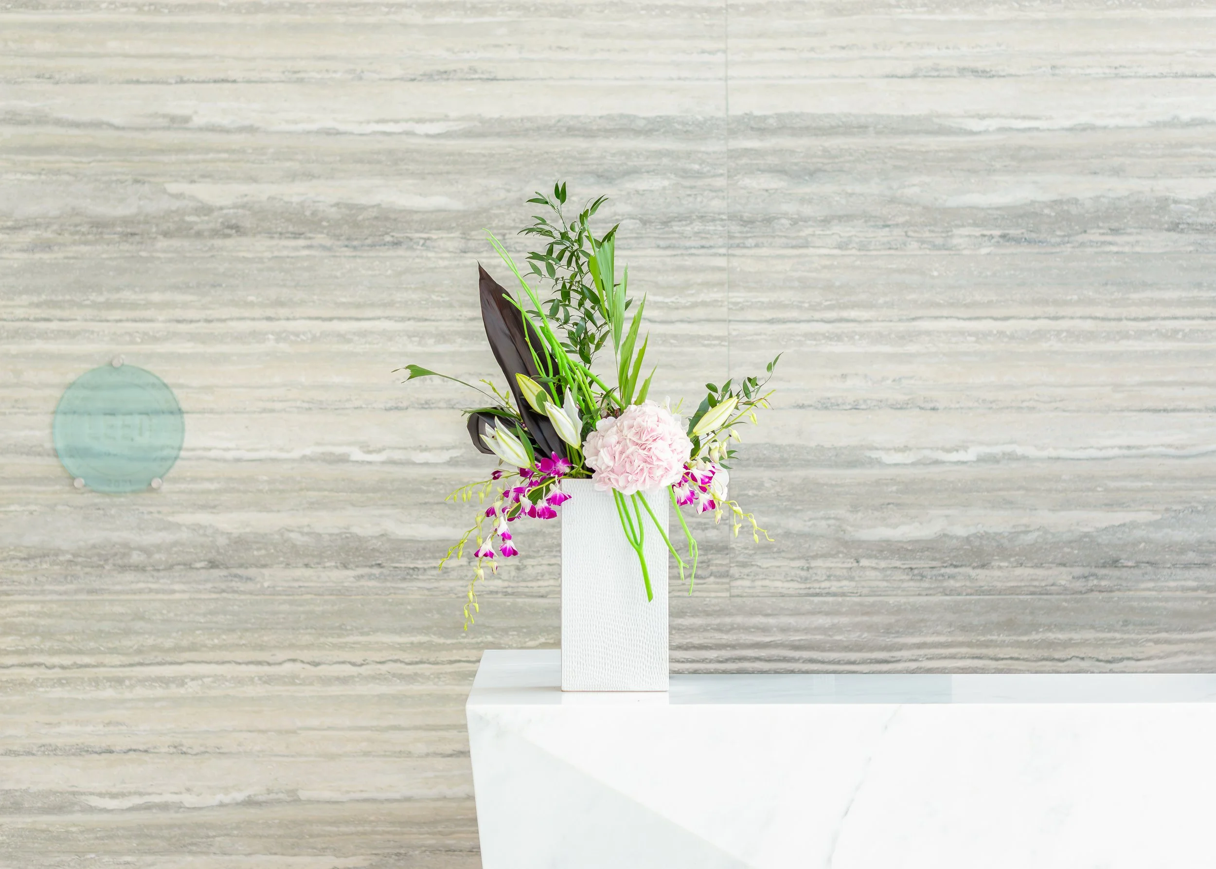 Elegant floral arrangement with pink flowers and green leaves in a white vase on a marble table against a textured gray wall.