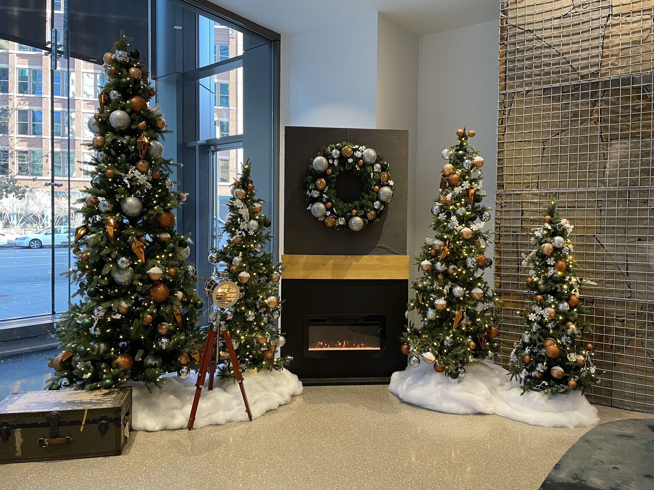 Christmas trees decorated with ornaments and lights, placed in a modern indoor setting with a fireplace and a wreath on the wall. Snow-like fabric covers the base of the trees, and there is a vintage suitcase and a spotlight tripod nearby.
