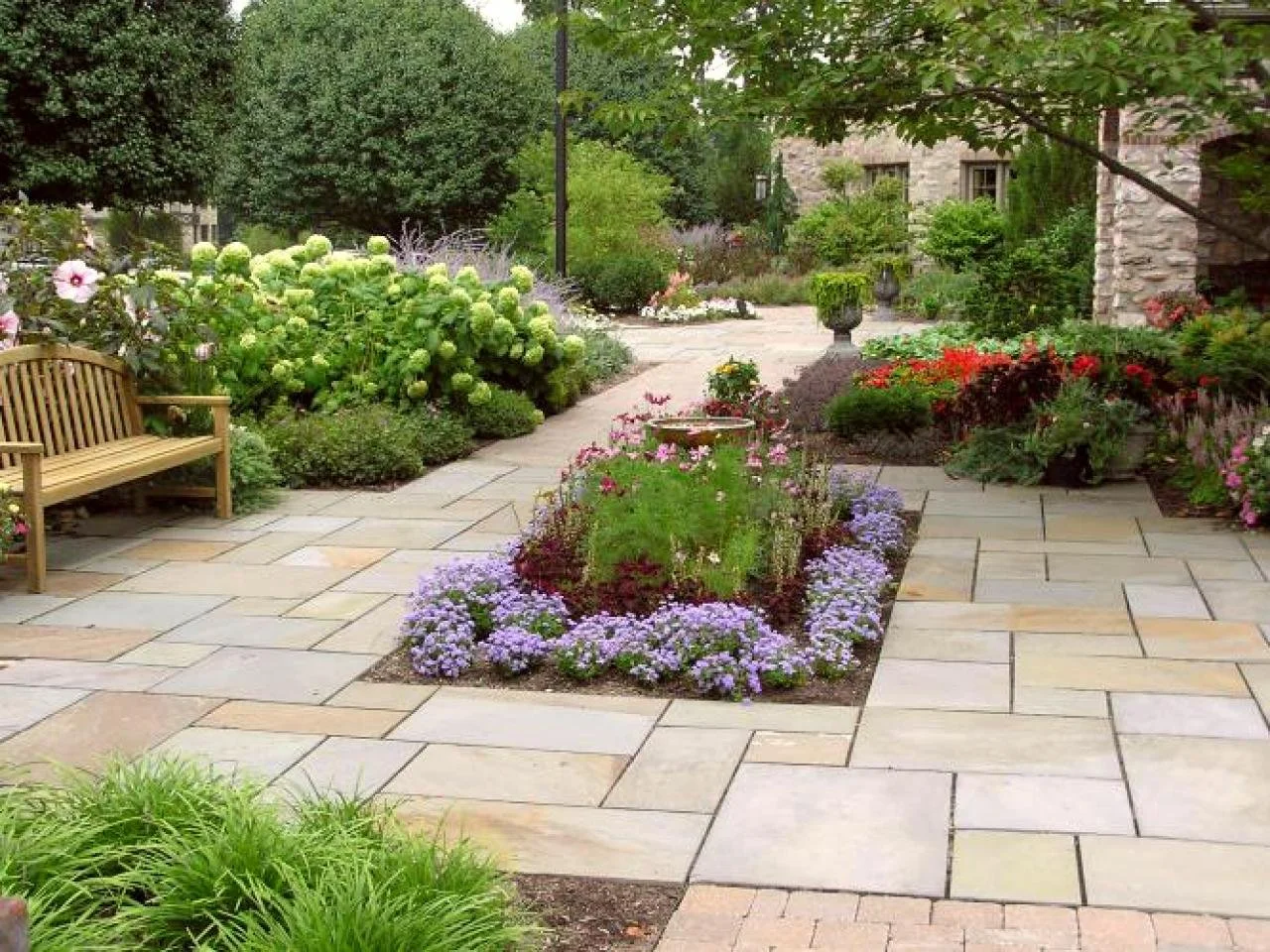 Garden scene with stone pathway, wooden bench, various flower beds, shrubs, and greenery.
