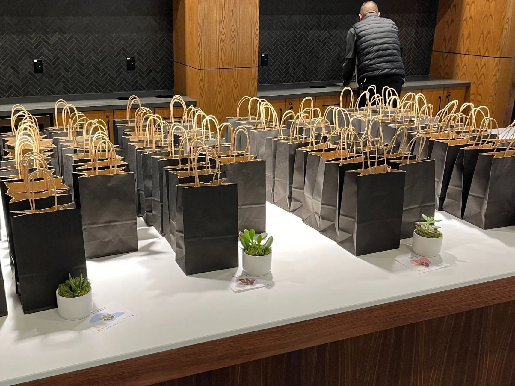 A table with numerous black gift bags, each with handles, arranged neatly. Small potted plants are placed at intervals on the table. A person in a black jacket stands in the background, facing away from the camera.