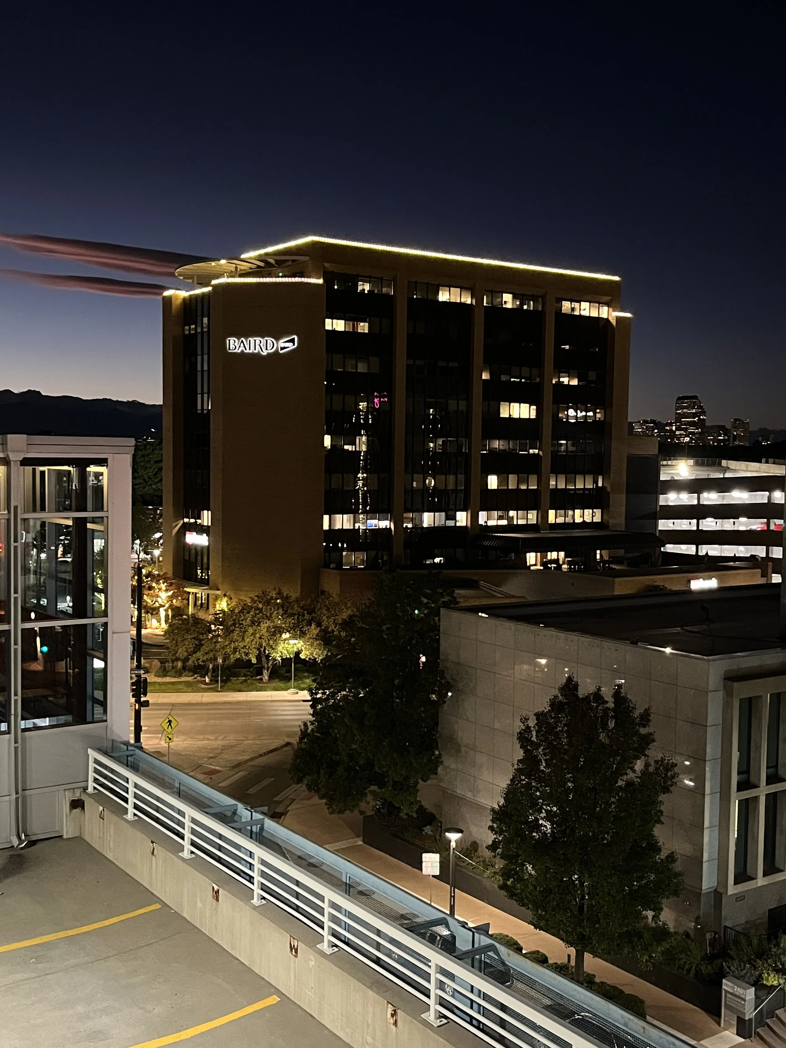 Night view of a well-lit office building labeled "BAIRD," with street and parking area, and a clear sky in the background.
