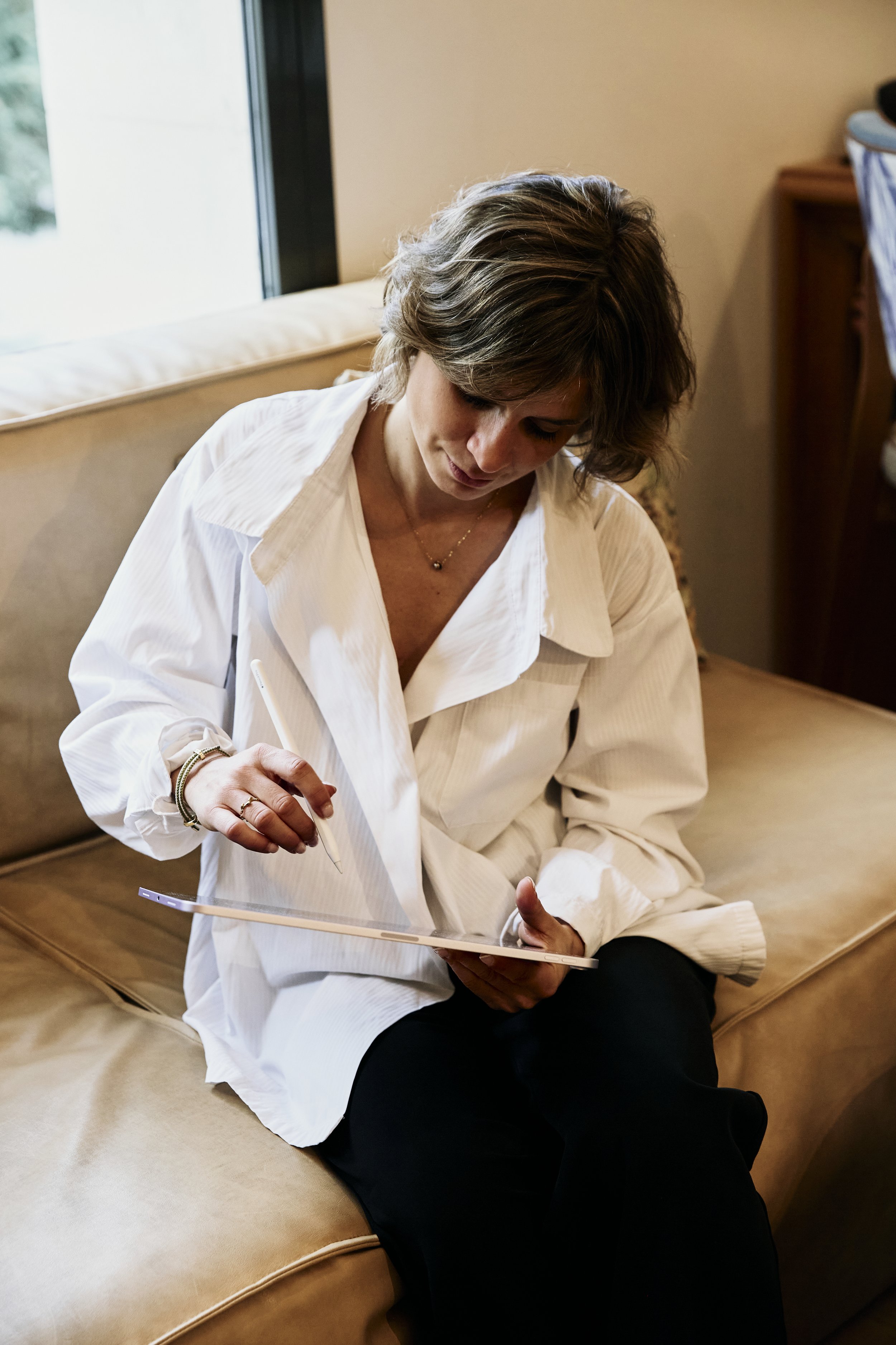A woman with short wavy hair wearing a white oversized shirt and black pants sitting on a beige sofa, looking down at a tablet device in her hands and using a stylus.